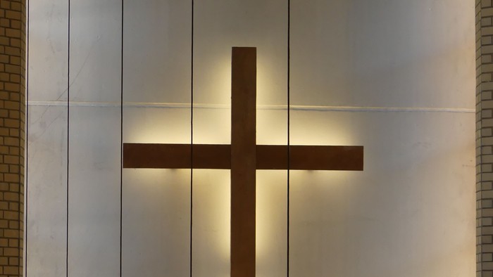 Moderne Metallkreuz, von Sonnenlicht beleuchtet, an einer reflektierenden Wand mit kerzenartigen Leuchten darunter.