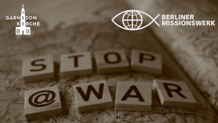 Eine friedensbezogene Botschaft „STOP @KRIEG“, gebildet aus Holzklötzen auf einem Weltkarten-Hintergrund mit Logos.