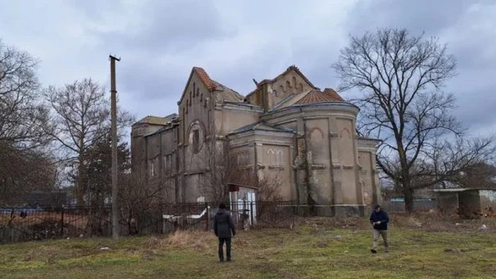 Eine beschädigte, verlassene Kirche steht in einer kargen Landschaft, während zwei Menschen sich dem Gebäude nähern.