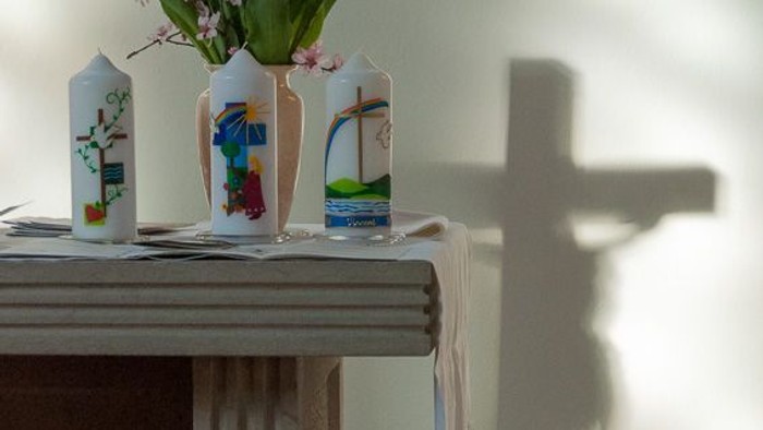 Verzierte Kerzen und Blumen auf einem Tisch mit einem Kreuz, das einen Schatten in einer ruhigen Innenraumeinstellung wirft.