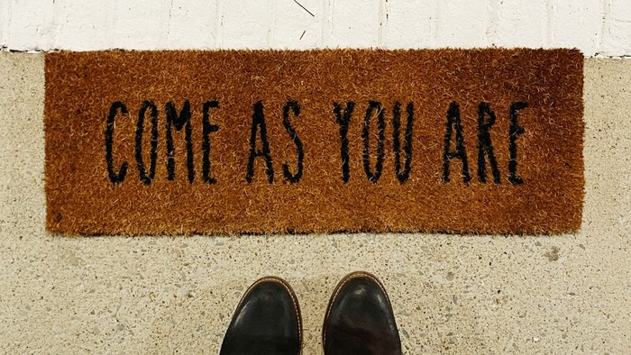 Eine Fußmatte mit der Aufschrift „Komm, wie du bist“ und schwarze Schuhe, die auf hellem Untergrund stehen.