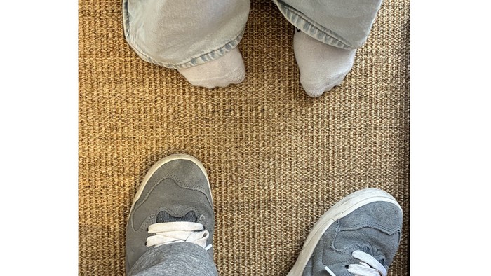 Fødder i lysegrå sneakers og jeans, der står på et vævet tæppe.