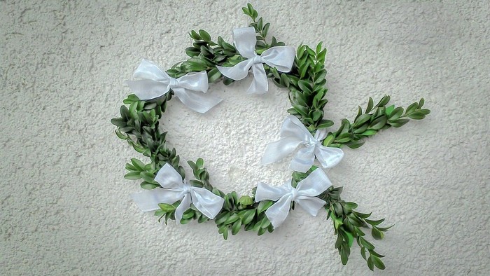 Ein Kranz aus grünen Blättern mit weißen Schleifen an einer weißen Wand.