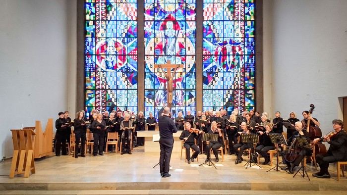 Ein Chor und Orchester, schwarz gekleidet, in einem Altarraum vor einem großen modernen Kirchenfenster