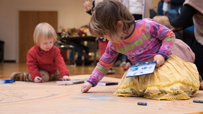 Zwei kleine Kinder malen kreativ und spielen mit Stiften auf einer großen Holzbodenfläche.