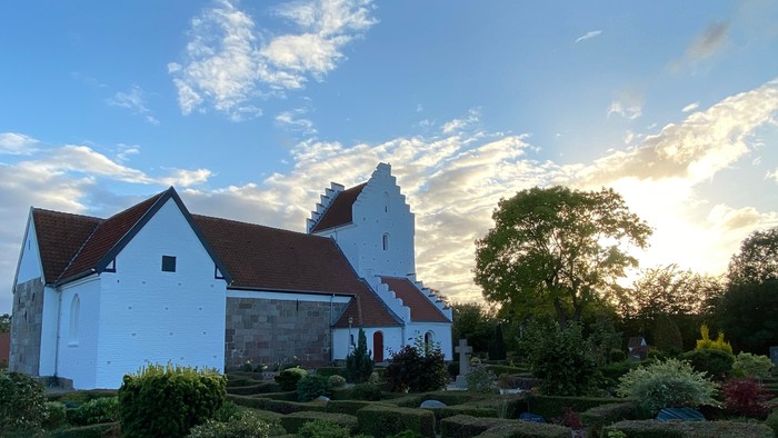 En hvid kirke med rødt tag og en have med grønne planter og træer ved solnedgang