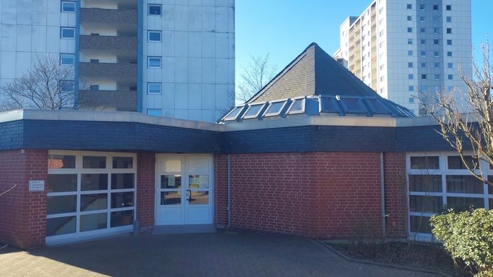 Kleines Ziegelgebäude mit Oberlichtern und großen Wohnhochhäusern im Hintergrund an einem klaren Tag.