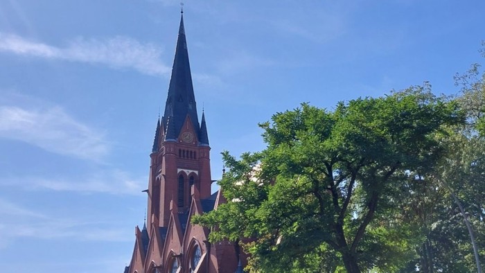 Ein roter Kirchturm mit spitzer Spitze vor grünen Bäumen und blauem Himmel