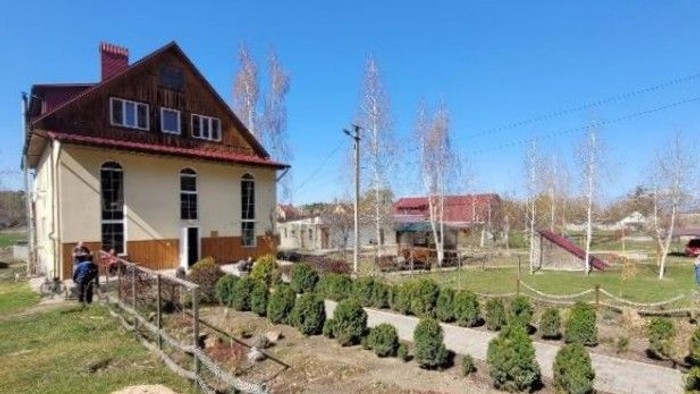 Ein kleines Haus mit Garten und Wege, umgeben von Bäumen und Himmel.