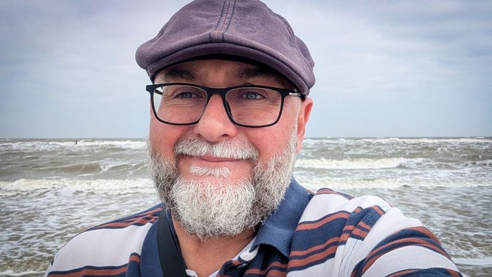 Ein Mann macht ein Selfie am Meer. Er trägt eine Schiebermütze und eine Brille und blickt mit einem leichten Lächeln in die Kamera. Sein grauer Bart ist deutlich zu sehen. Im Hintergrund erstreckt sich eine bewegte Wasserfläche mit Wellen unter einem bewö