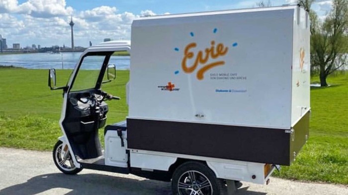 Elektrisches Lieferdreirad mit der Marke "Evie" für nachhaltige Paketzustellung in der Stadt in der Nähe einer malerischen Uferpromenade.