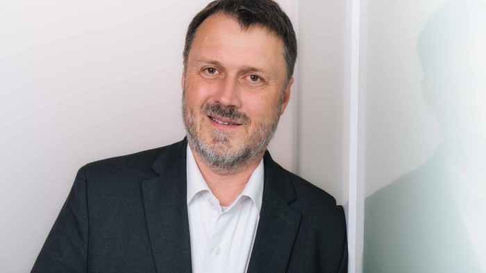 Professioneller Mann im Anzug, der vor einem minimalistischen Hintergrund in die Kamera lächelt.