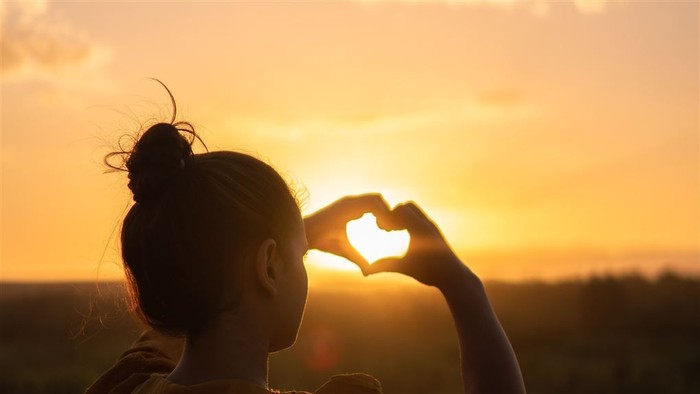 En person med hår i en bun holder hænderne i hjerteform foran solnedgang