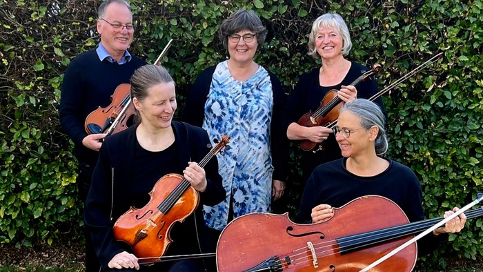 Eine Gruppe von fünf Musikern posiert mit ihren Streichinstrumenten vor einem grünen Busch.