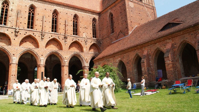 Mönche in weißen Roben stehen in einem historischen Kreuzganghof mit gewölbter Backsteinarchitektur.