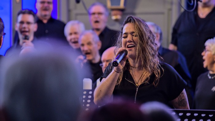 Ein Chor tritt mit einer Leadsängerin/einem Leadsänger auf, die/der während eines Konzerts leidenschaftlich in ein Mikrofon singt.