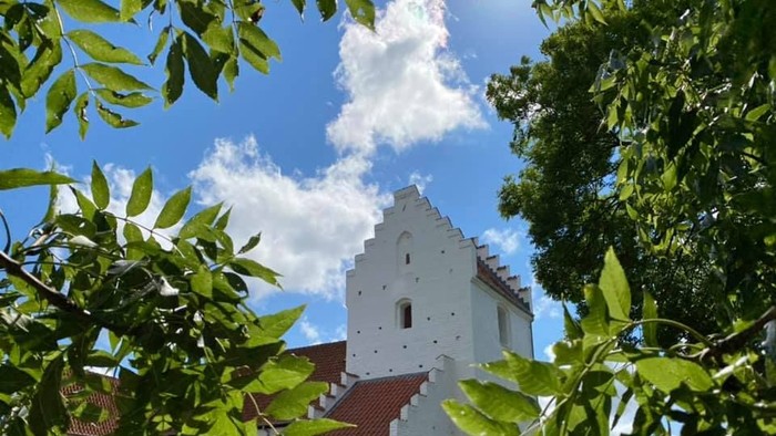 Et hvidt kirkebygning med røde tag og en tårn, omgivet af grønne træer og buske