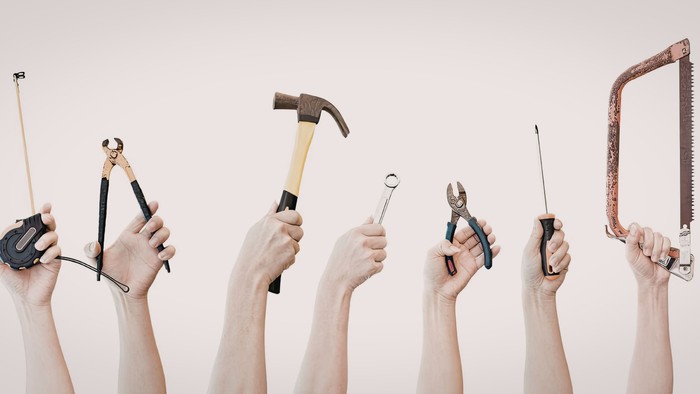 Samling af hænder, der holder forskellige essentielle håndværktøjer som hammer, tang, sav og skruetrækker.