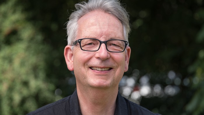 Ein lächelnder Mann mit Brille und grauen Haaren, der ein dunkles Hemd trägt, im Freien.