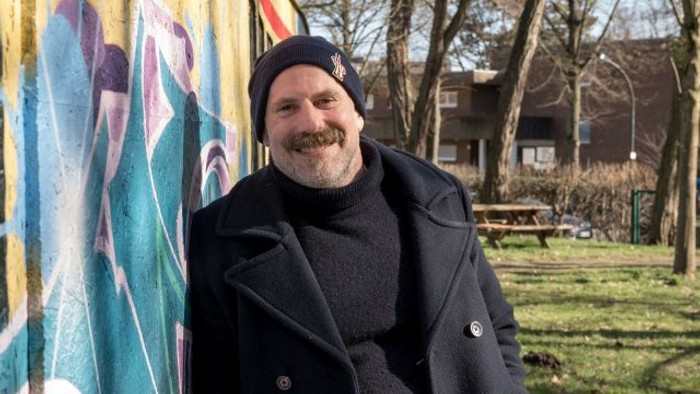 Ein lächelnder Mann in einem Mantel steht neben einem bunten Wandgemälde in einer parkähnlichen Umgebung.