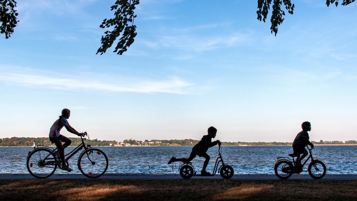 Kinder fahren auf Fahrädern und Roller an einem See entlang.