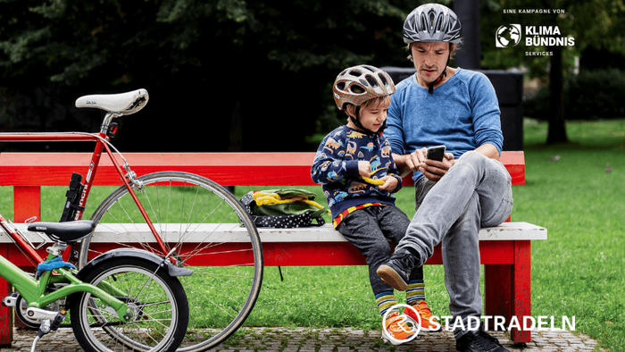 Auf einer roten Bank in einem Park sitzen ein Vater und sein Sohn. Beide tragen Fahrradhelme. Ihre Fahrräder haben sie an der Bank angelehnt. Das Bild wirbt für die Aktion „STADTRADELN”. Das Logo ist am Rand des Bildes zu sehen.