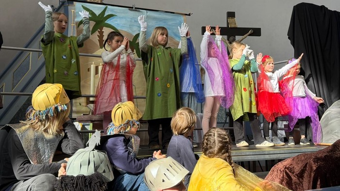 Kinder führen in einer Kirche ein Ostermusical auf: Eine Gruppe steht auf einer kleinen Bühne vor einem Kreuz und einer bemalten Kulisse mit Landschaft. Die Kinder tragen bunte Kostüme mit Tüchern und heben synchron die Arme zu einer Choreografie. Im Vord