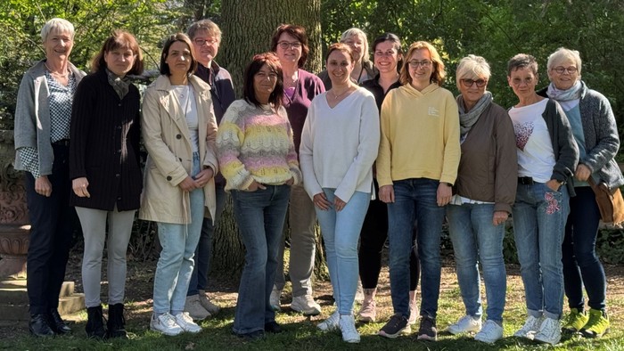 Gruppe von etwa zwölf Frauen steht im Freien vor einem großen Baum in einem parkähnlichen Garten. Die Frauen sind unterschiedlich alt, tragen Alltagskleidung und stehen dicht nebeneinander, lächelnd in die Kamera. Im Hintergrund sind viel Grün, Sträucher 