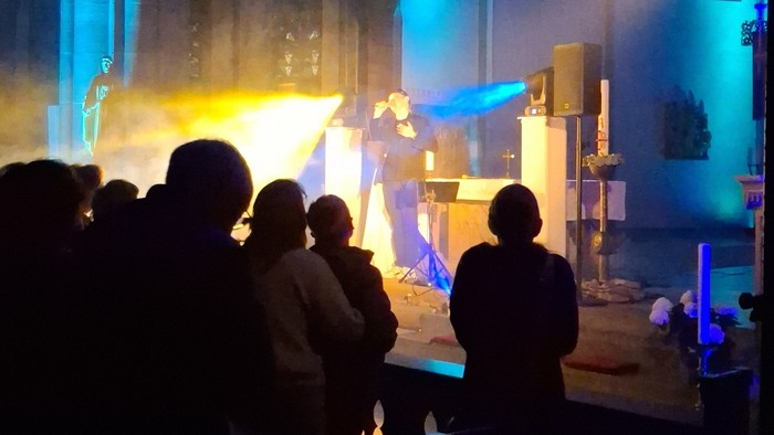 Eine Menschenmenge beobachtet eine dramatische Bühnenaufführung mit blauer Beleuchtung und einem hellen Scheinwerfereffekt.