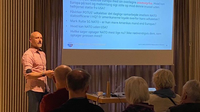 En taler stiller NATO-relaterede spørgsmål til et opmærksomt publikum under en diskussion.