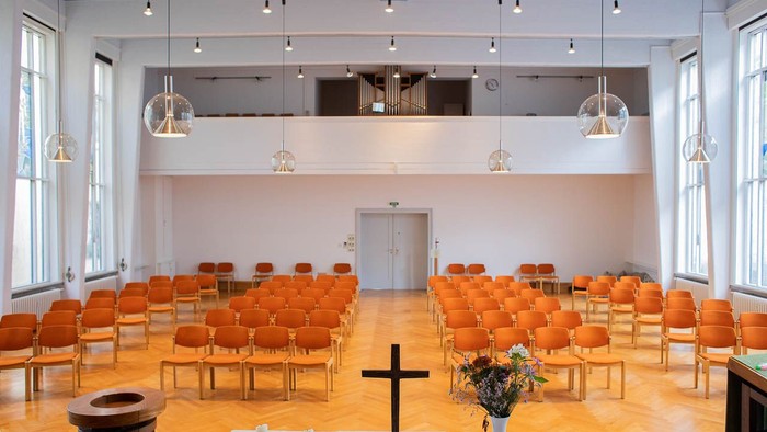 Moderne Kapelle mit orangefarbenen Stühlen, Holzboden und einem schlichten Altar mit Kreuz und Blumenschmuck.