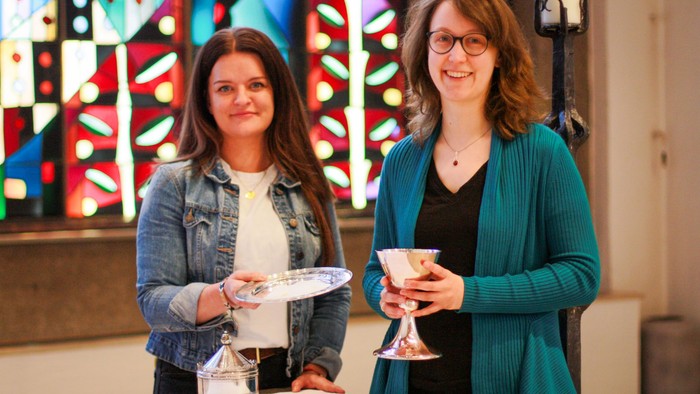 Zwei junge Frauen, Miriam Attila und Pfarrerin Theodora Beer, zeigen Abendmahlsgerät und Oblaten