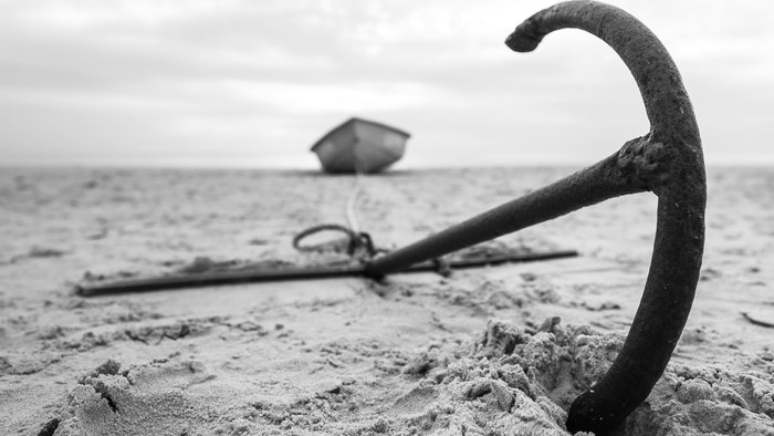 Verankerter Anker, halb im sandigen Strand vergraben, mit einem fernen Boot im Wasser.