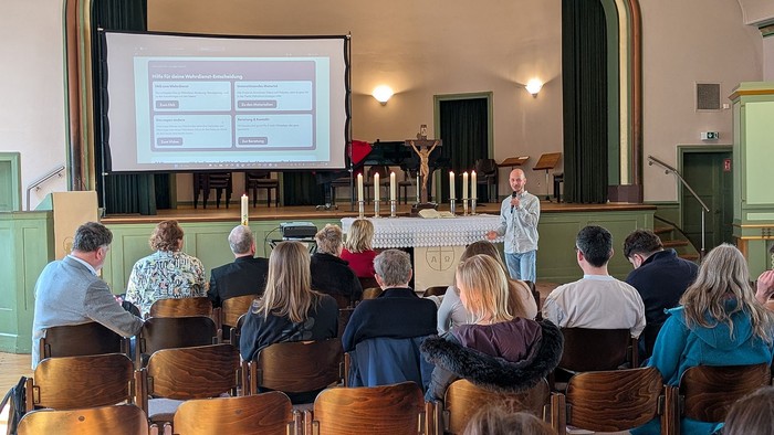 Ein Referent steht vorne in einem Gemeindesaal vor Altar und Kerzen und spricht mit Mikrofon zu einer Gruppe von Zuhörenden, die auf Stuhlreihen sitzen. Hinter ihm ist eine große Leinwand mit einer Präsentation zum Thema Wehrdienst-Entscheidung zu sehen.