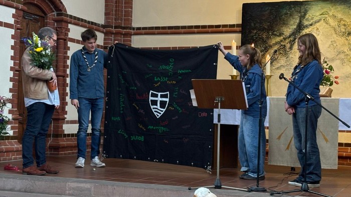 Drei Jugendliche halten vor dem Altar ein großes schwarzes Tuch mit bunten Namen und einem weißen Symbol hoch.
