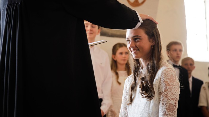 En ung pige i hvidt kjole smiler og ser op, mens hun står ved et mørkt tæppe.