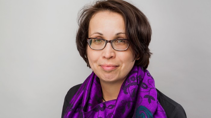 Eine Person mit Brille und einem lebhaften lila gemusterten Schal lächelt sanft vor einem neutralen Hintergrund.