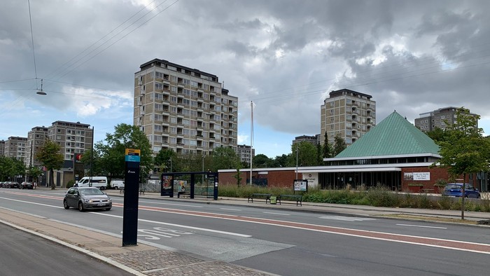 Bymæssig gadescene med højhuse, en busstoppested og en nærliggende struktur med grønt tag.