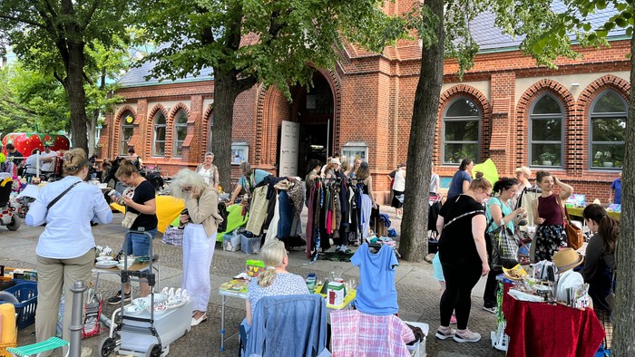Belebter Freiluftmarkt mit Menschen, die zwischen verschiedenen Waren und Ständen unter schattigen Bäumen stöbern.