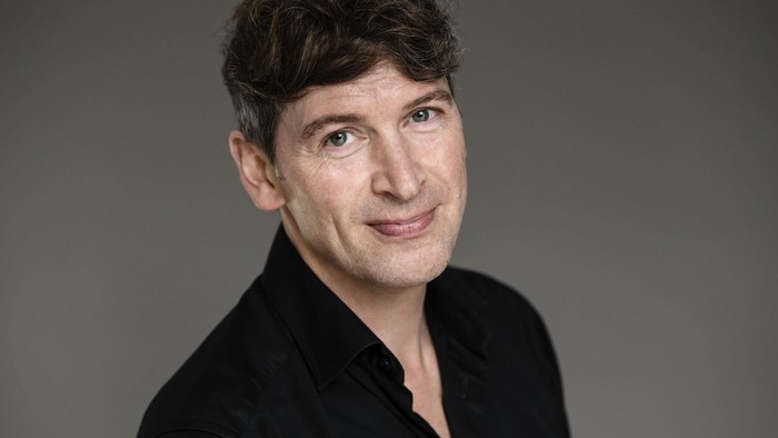 En mand med kort, mørkt hår smiler mildt i en sort skjorte på en neutral baggrund.