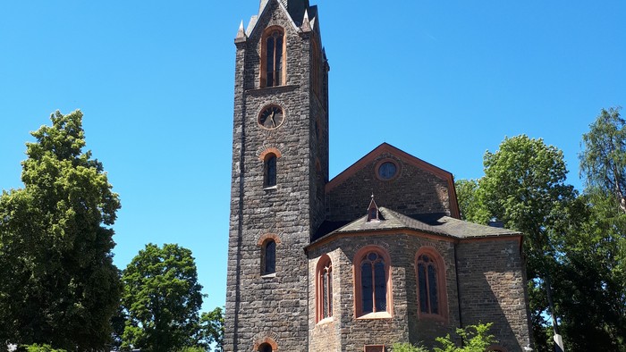 Historische Steinkirche mit hohem Turm, umgeben von üppigem Grün unter klarem blauem Himmel.