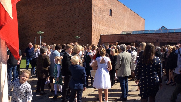 En folkemængde er samlet foran et rødt murstenshus med blå tag.