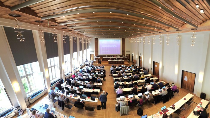 Großer Hörsaal, gefüllt mit sitzenden Zuhörern, die einer Präsentation auf einer Leinwand vorne folgen.