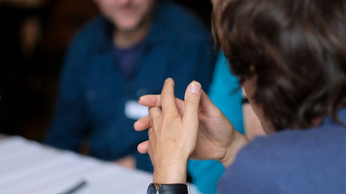 Drei Menschen sitzen zum Gespräch zusammen. Im Fokus ist ein Mann auf der rechten Seite, welcher von hinten mit verschränkten Fingern zu sehen ist.