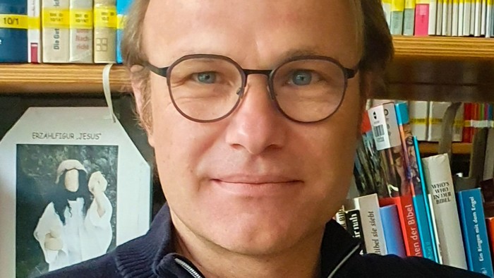 Ein Mann mit Brille und einem hohen blonden Scheitel vor einem Bücherregal