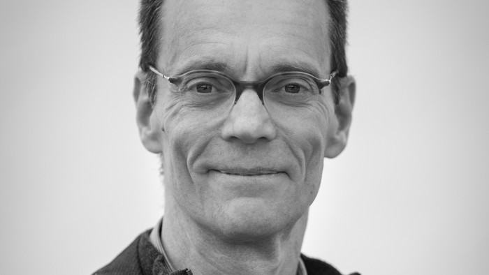 Ein lächelnder Mann mit Brille, der einen Blazer und einen Pullover trägt, posiert vor einem schlichten Hintergrund.