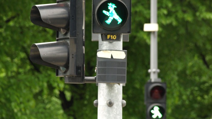 Fußgängerampel mit grünem Geh-Symbol an einem Pfosten, dahinter grüne Bäume.