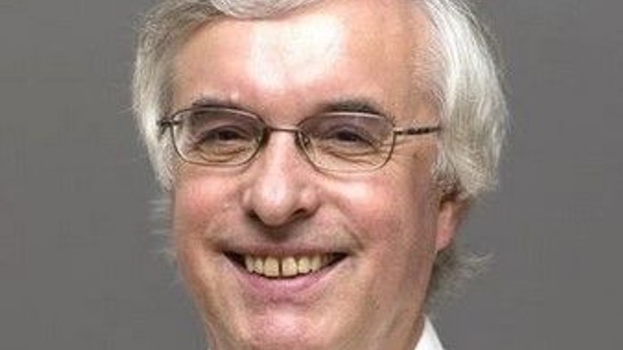 Ein lächelnder Mann mit weißem Haar, Brille und einem hellen Hemd mit Knöpfen.