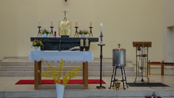 Ein ruhiger Kirchenaltar, geschmückt mit Kerzen, Blumen, religiösen Gegenständen und einem Taufbecken.