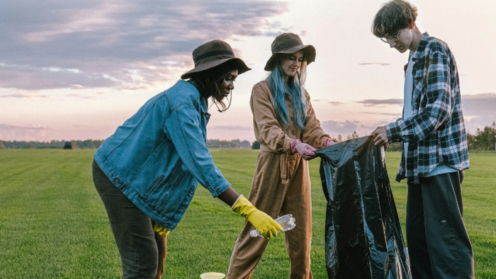 Drei Personen mit Handschuhen sammeln Müll auf einem grünen Feld während des Sonnenuntergangs.